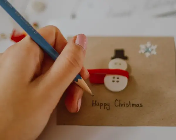 Ręka z ołówkiem pisze "Happy Christmas" na kartce z bałwankiem z guzików. Idealny przykład, jak narysować kartkę świąteczną.