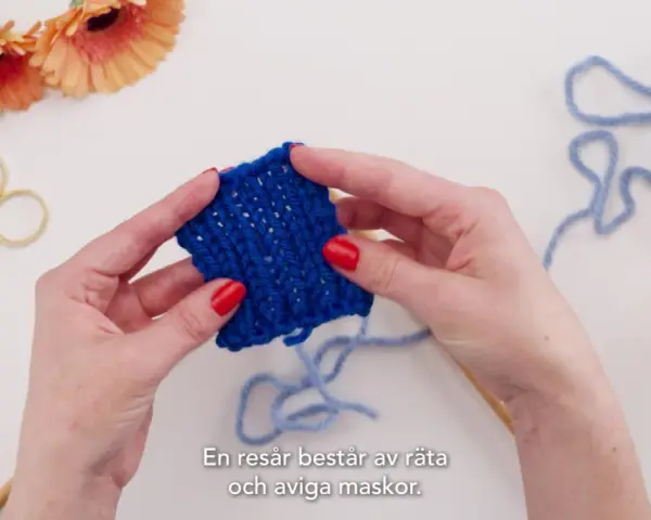 Händer som håller i ett stickat provstycke i resårstickning. Blått garn, sax och blommor syns i bakgrunden.