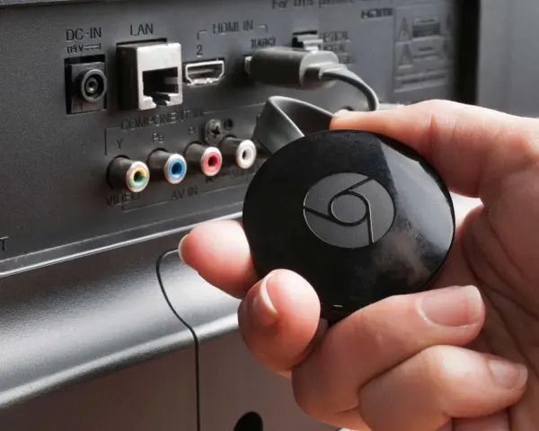 En hand håller en svart Chromecast för att ansluta till en HDMI-port på baksidan av en TV.