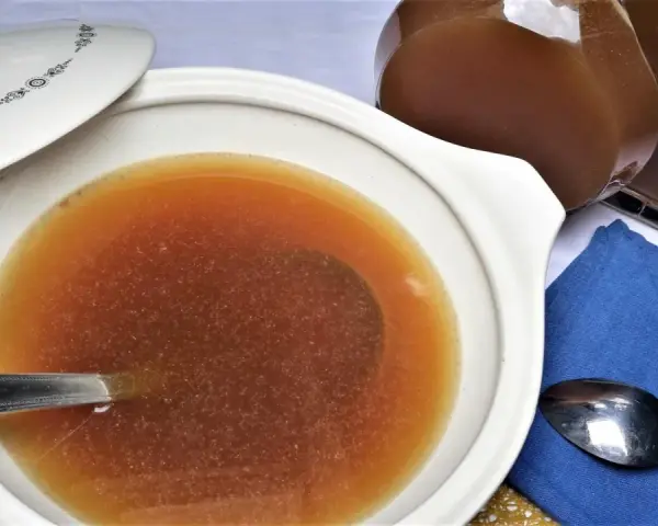 Caldo de carne casero en una cazuela blanca con tapa, junto a una jarra y cucharas.