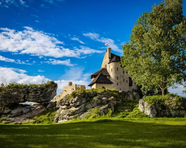 Zamek Bobolice na skale, z polską flagą, pod błękitnym niebem z chmurami. Obok skalny most i zielona trawa.