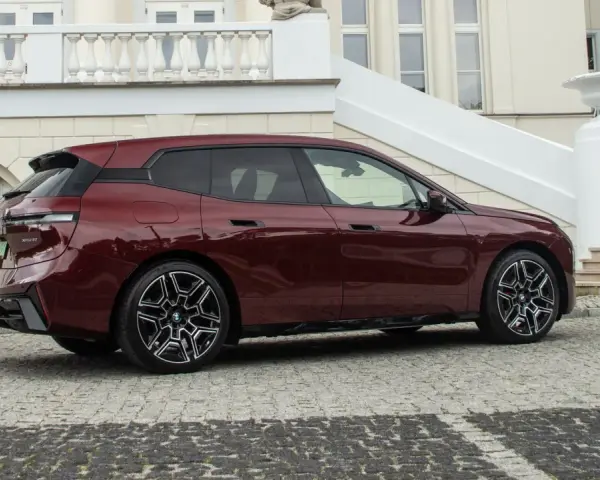 Ciemnoczerwone BMW iX stoi na brukowanym podjeździe przed eleganckim budynkiem.