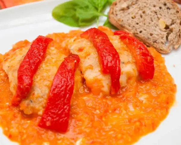 Bacalao a la riojana, un plato tradicional con trozos de pescado blanco cubiertos de pimientos rojos y una salsa de tomate y pimientos. Servido con pan y hojas verdes.