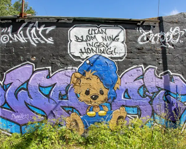 Enkel graffiti på en tegelvägg visar en brun nallebjörn i blå overall och mössa, med texten "Utan blommor ingen honung!".