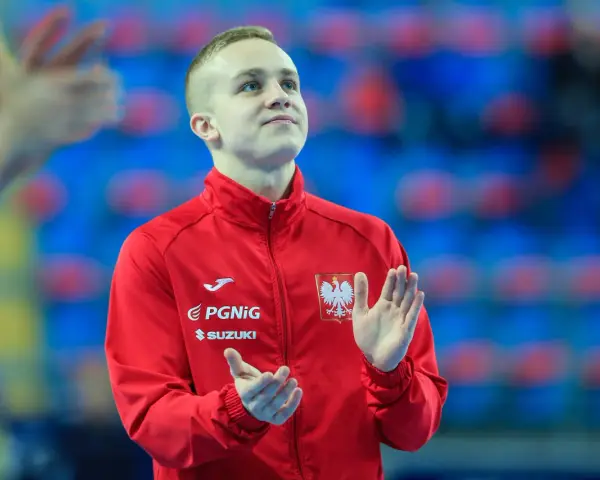 Adam Jędraszek w czerwonej kurtce z orłem, z dłońmi uniesionymi w geście radości lub powitania.