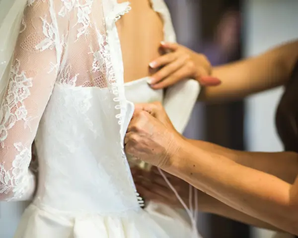 Dettaglio di abiti da sposa riutilizzabili: mani che chiudono bottoncini su un vestito bianco con pizzo e velo.