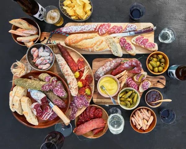 Eine reichhaltige Wurstplatte anrichten mit Baguette, Oliven, Cornichons und Senf.
