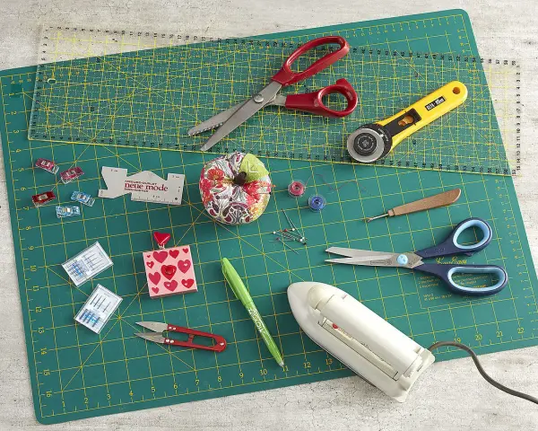 Matériel de couture : ciseaux, cutter rotatif, aiguilles, épingles, fils, fer à repasser, sur un tapis de découpe.