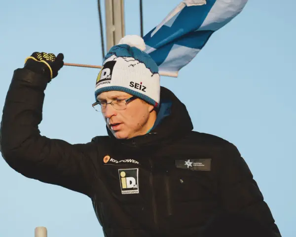 Igor Medved w czapce z pomponem i flagą Finlandii, w czarnej kurtce z logo.