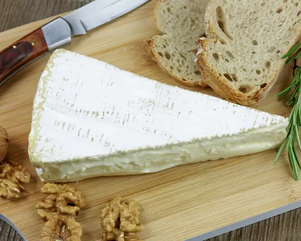 Tranche de fromage à croute lavée crémeux, accompagnée de pain, noix et herbes aromatiques sur une planche en bois.