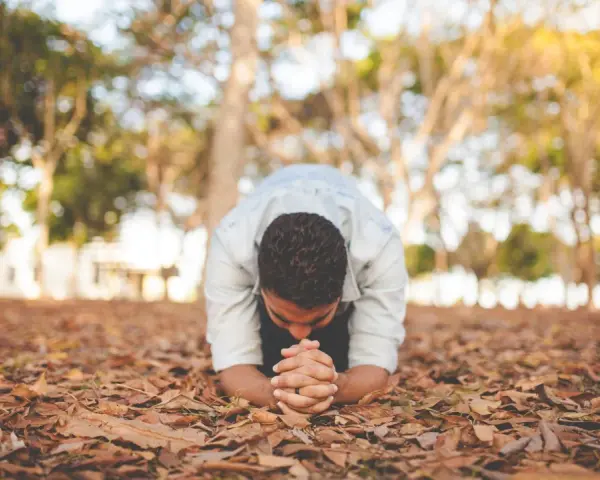 A man in a light blue shirt is in a posture of christian prostration, hands clasped on the ground covered in fallen leaves.