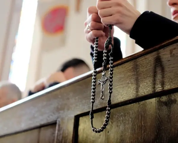 Dłonie trzymające różaniec podczas nabożeństwa. W tle widać innych wiernych, przygotowujących się na odpust.