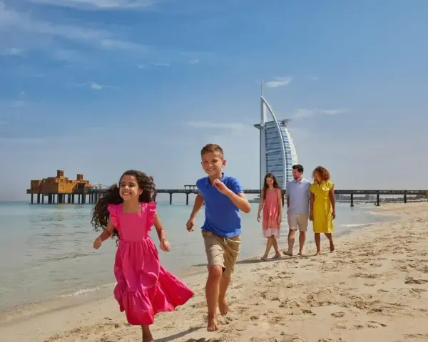 Rodzina cieszy się słonecznym dniem na plaży w Dubaju. Dzieci biegną w stronę morza, a w tle widać słynny hotel Burj Al Arab.