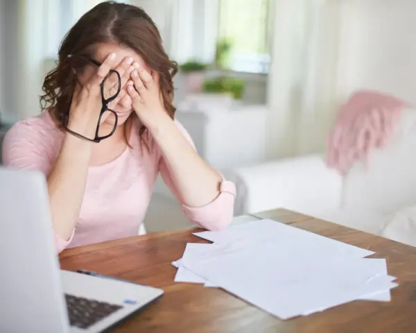 Kobieta z okularami w dłoniach, zamyka oczy, czując stres przed nową pracą. Na stole laptop i dokumenty.