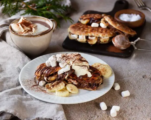 En härlig nutella macka med banan och marshmallows, toppad med grädde och kakao. Bredvid står en kopp varm choklad.