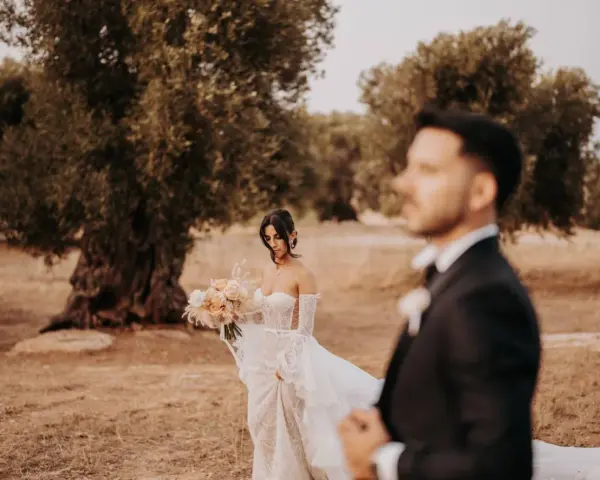 Sposarsi a maggio sotto un ulivo secolare. La sposa con un bouquet di rose pesca, lo sposo in primo piano sfocato.