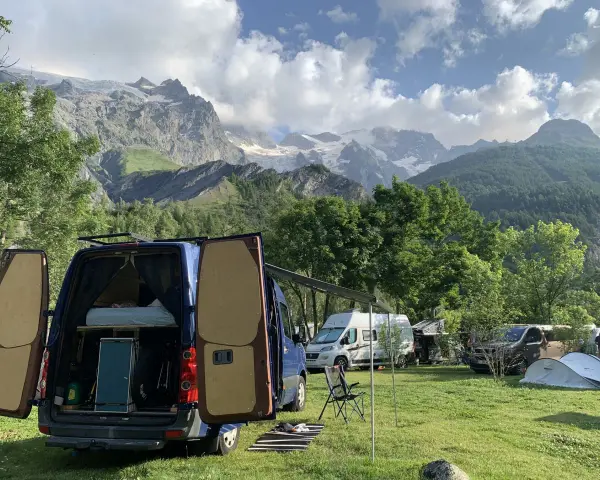 En campingplats med flera husbilar, inklusive en blå med öppna bakdörrar, i en dal med snöklädda berg i bakgrunden. En perfekt plats för äventyr med edins husbilar.