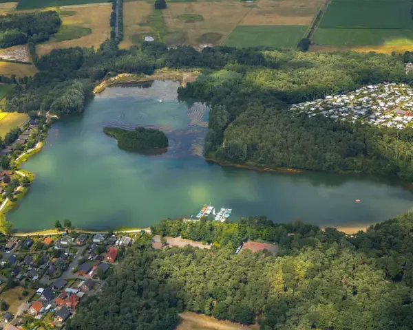 Luftaufnahme des Feldmarksees Sassenberg mit einer kleinen Insel, Booten am Steg, einem Wohngebiet und Feldern.