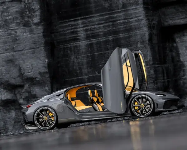 Szary Koenigsegg Gemera, czteromiejscny supersamochód z otwartymi drzwiami, prezentuje swoje żółte wnętrze na tle skalistej ściany.