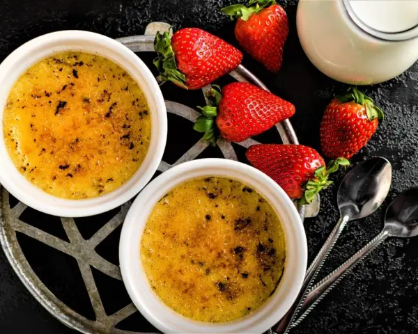 Zwei cremige Crème brûlée mit karamellisierter Kruste, garniert mit frischen Erdbeeren und einem Milchkännchen.