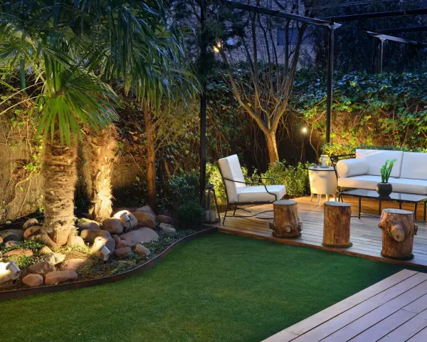 Un jardín poco mantenimiento con césped artificial, palmeras iluminadas y una zona lounge con sofá blanco y sillón.