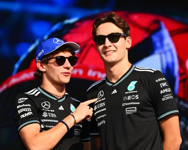 Dos pilotos de Fórmula 1, George Russell y un compañero, sonríen con gafas de sol. Russell lleva una gorra azul y ambos visten camisetas negras con logotipos de patrocinadores.