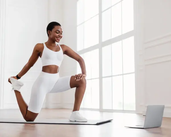 Femme en tenue de sport blanche effectuant un exercice quadriceps sur un tapis, guidée par un ordinateur portable.