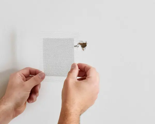 Hands apply a mesh patch to a hole in a white wall, demonstrating how to patch a hole in the wall.