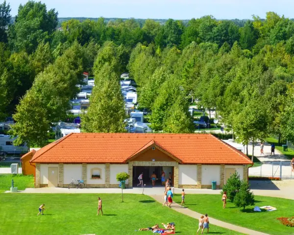 Słoneczny dzień na kempingu Liptot. Ludzie odpoczywają na trawie, a w tle widać rzędy kamperów i przyczep kempingowych wśród drzew.