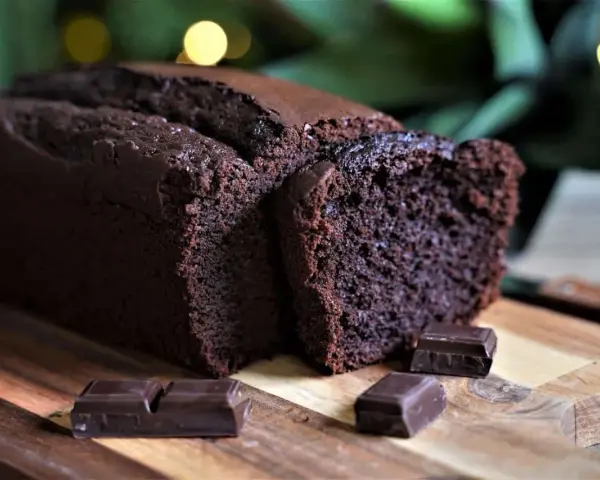 Un délicieux cake au chocolat, moelleux et intense, coupé en tranches sur une planche en bois, avec des carrés de chocolat noir en garniture.