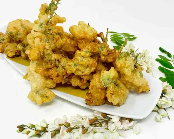 Fiori di acacia fritti, dorati e croccanti, serviti su un piatto bianco con rametti di fiori e foglie fresche.