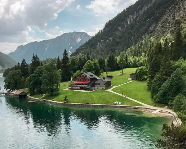 Gaisalm am Achensee: ein Gasthaus mit roten Fensterläden, umgeben von grünen Wiesen und Bergen. Ein Schiff liegt am Steg.