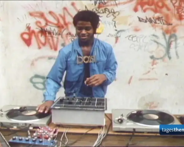 DJ Kool Herc, der oft als derjenige gilt, der Hip Hop erfunden hat, legt auf. Graffiti zieren die Wand im Hintergrund.