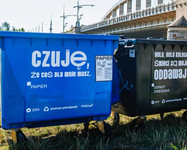 Niebieski kontener na makulaturę z napisem "czuję, że coś dla mnie masz. PAPIER" obok czarnego na odpady zmieszane.