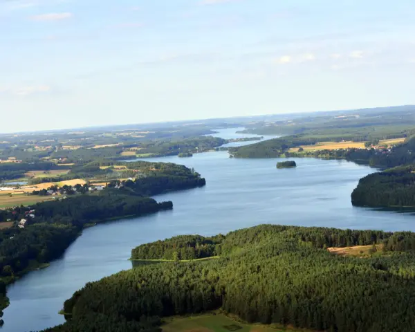 Rozległe jezioro raduńskie dolne otoczone lasami i polami. W oddali widać zabudowania.