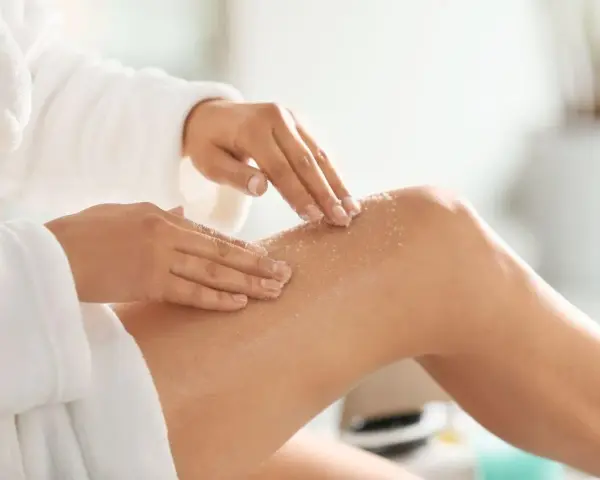 Kobieta w białym szlafroku wykonuje peeling nóg. To idealny peeling przed goleniem, by skóra była gładka.