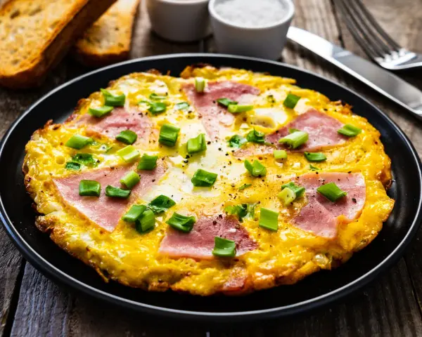 Pyszny omlet z szynką i serem, posypany szczypiorkiem, podany z grzankami. Idealny na śniadanie.