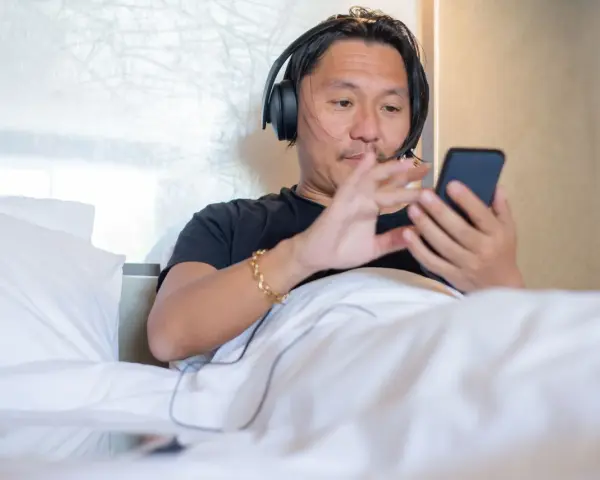 Mężczyzna w słuchawkach leży w łóżku, korzystając z telefonu. To prosty sposób, jak słuchać audiobooka na telefonie, relaksując się.