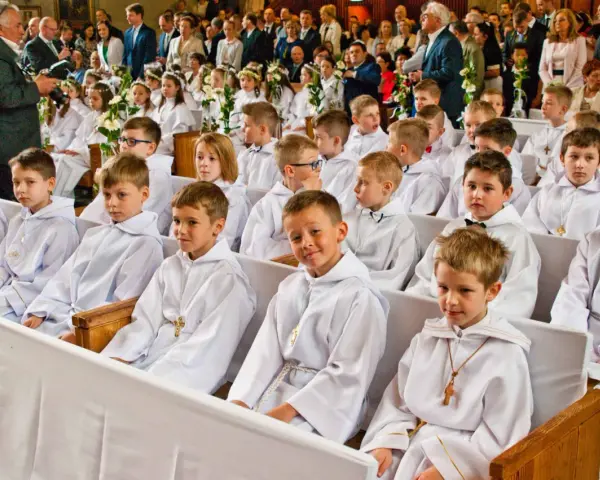 Dzieci w bieli, gotowe na rocznicę komunii świętej, siedzą w kościele. W tle widać zgromadzonych gości i fotografa.