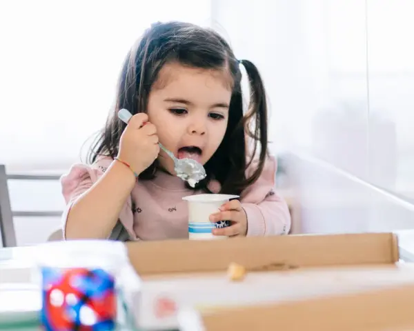 Mała dziewczynka z kitkami je jogurt z kubeczka. Od kiedy dziecko może jeść danonki? Odpowiedź jest prosta: gdy tylko zaczyna jeść pokarmy stałe.