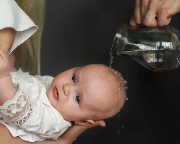 Chrzest dziecka. Woda spływa po głowie malucha w białej szatce. Niech to będzie początek mądrych życzeń na chrzest.