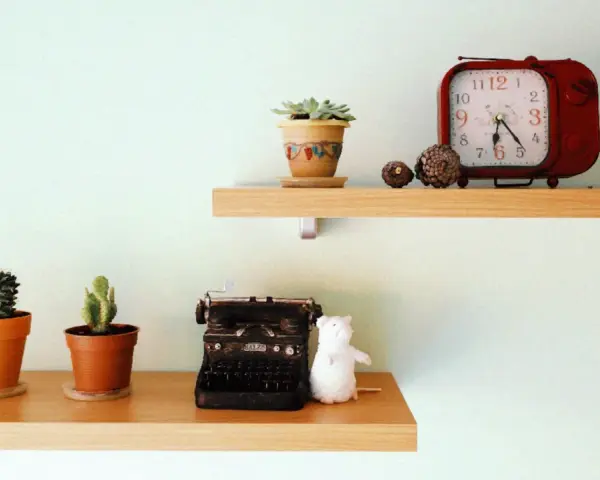 Ideas para decorar baldas salón: dos estantes de madera con cactus, una máquina de escribir antigua, un peluche blanco y un reloj rojo.