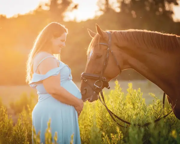 Une femme enceinte en robe bleue caresse un cheval brun au coucher du soleil. L'équitation enceinte est une belle façon de célébrer la maternité.