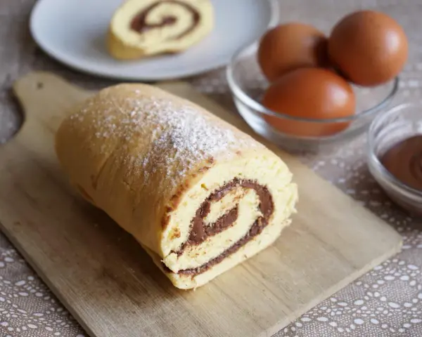 Un delizioso rotolo alla nutella, spolverato di zucchero a velo, pronto per essere gustato. Ingredienti freschi, uova e crema al cioccolato, completano la scena.