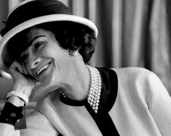 Coco Chanel sorride con un cappello e perle, un'icona di stile che sembra raccontare la storia di quando è nata Coco Chanel.