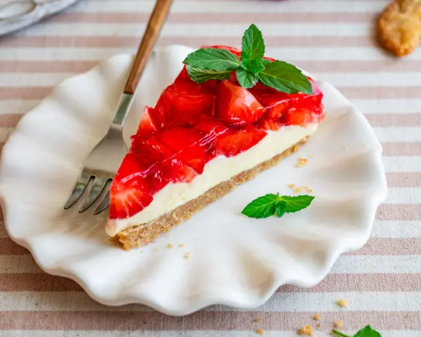 Ein Stück Erdbeerkuchen ohne Backen, mit frischen Erdbeeren und Minze garniert, auf einem weißen Teller.
