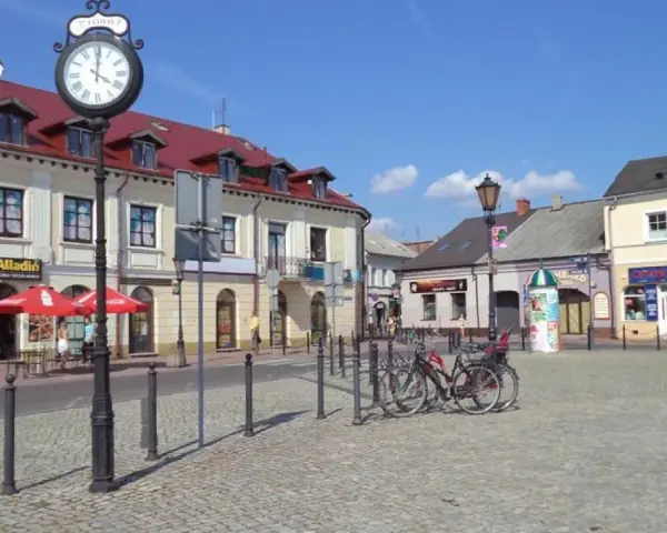 Słoneczny stary rynek łowicz z brukowanym placem, budynkami z czerwonymi dachami i zegarem na słupie.