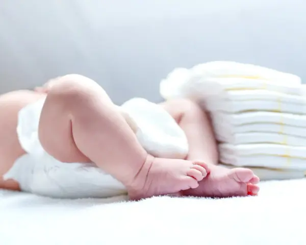 Niemowlę w pieluszce obok stosu nowych. Ile pieluszek powinno moczyć niemowlę? To zależy od wielu czynników.