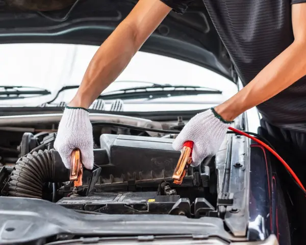 Hands in white gloves connect jumper cables to a car battery. This process helps answer how long does it take to charge a car battery.