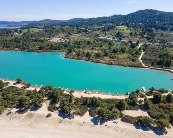 Turkusowa laguna na Kassandrze, otoczona zielonymi wzgórzami i lasem sosnowym. W oddali widać zabudowania i plażę z łodziami.